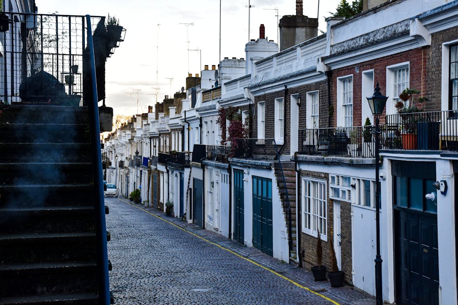 A row of residential terraced houses in Ilford with brick facades and white window frames, featuring external metal staircases painted in blue leading to first-floor entrances. Some staircases have ha