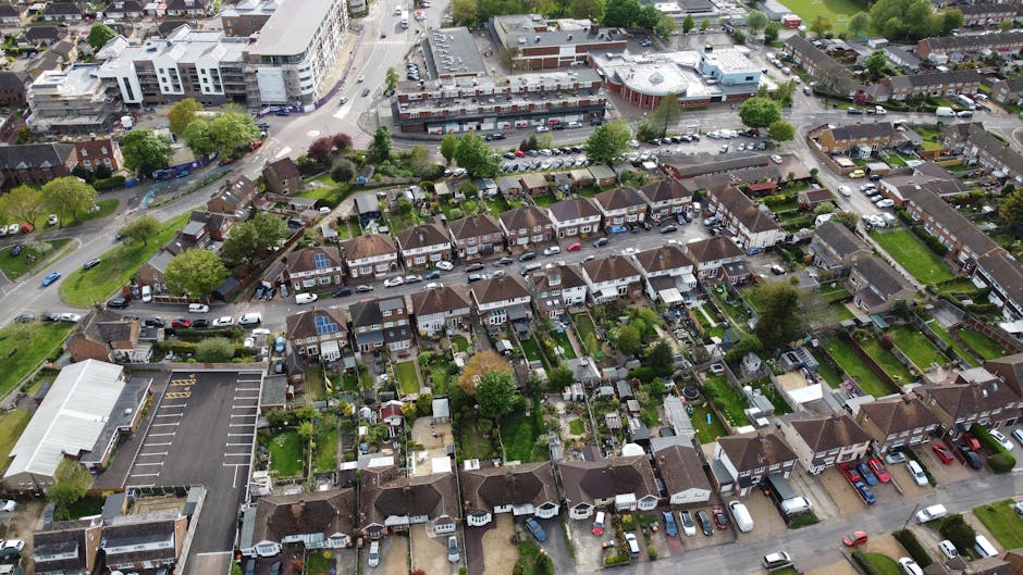 A residential street scene featuring parked cars lining both sides of the street, with a variety of vehicles including small hatchbacks, larger vans, and a covered item on a trolley. The pavement runn